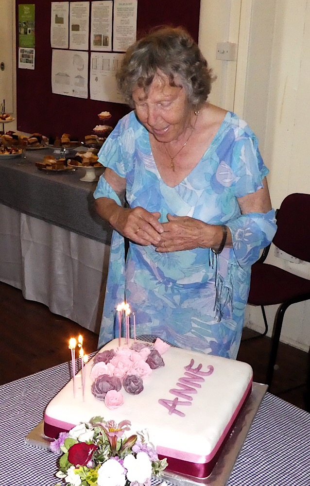 A picture of Anne Clark, wearing a blue dress and matching scarf, looking at her birthday cake